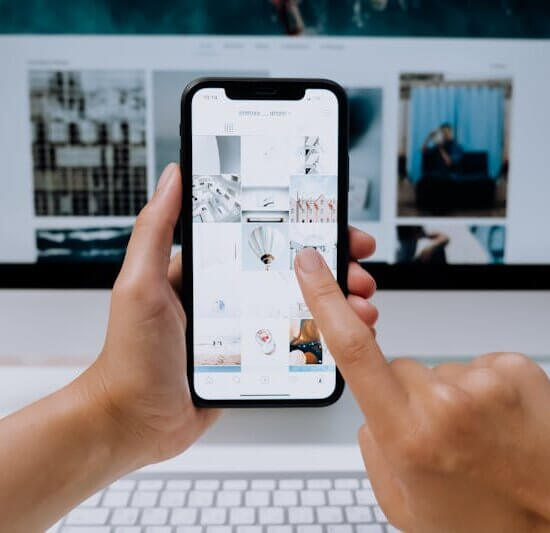 Hands holding a smartphone while browsing a social media app in front of a desktop computer.